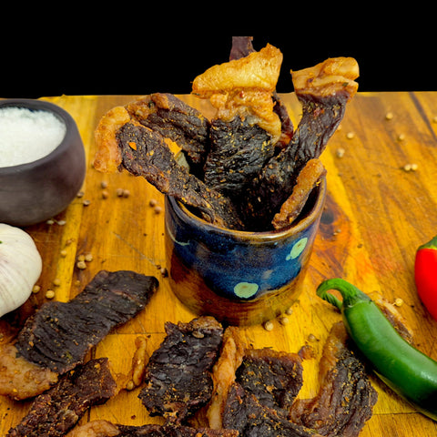 Dried beef biltong pieces in a blue clay pot on a wooden cutting board with spices, garlic and chili.