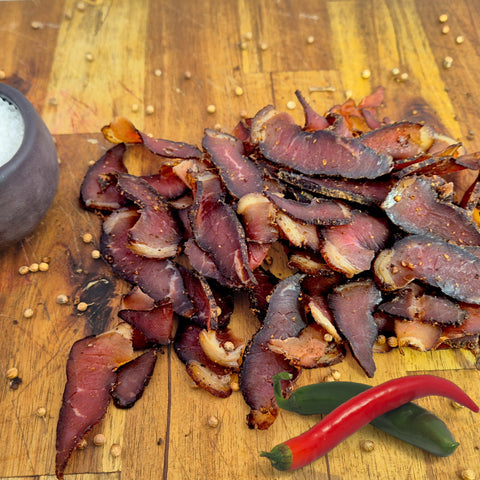 Biltong beef slices on a wooden cutting board with a red chili pepper and a salt pot.